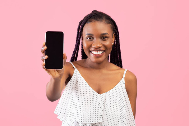 Positive African American lady holding cellphone with mockup for design, smiling at camera over pink studio background - Фото, изображение