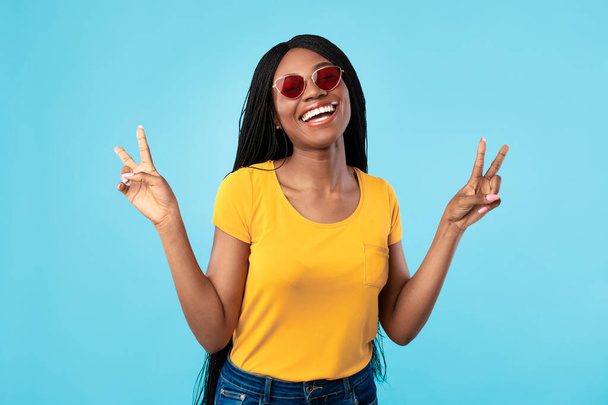 Positive afroamerikanische Frau gestikuliert V-Zeichen trägt Sonnenbrille, blauer Hintergrund - Foto, Bild