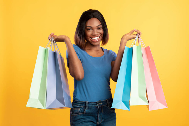 Vreugdevolle Afrikaanse Amerikaanse vrouw met kleurrijke shopper tassen, gele achtergrond - Foto, afbeelding