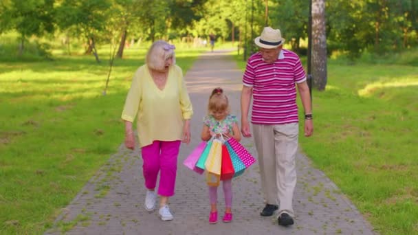 Niña niña caminando con el abuelo jubilado mayor abuela con bolsas de colores después de ir de compras - Metraje, vídeo