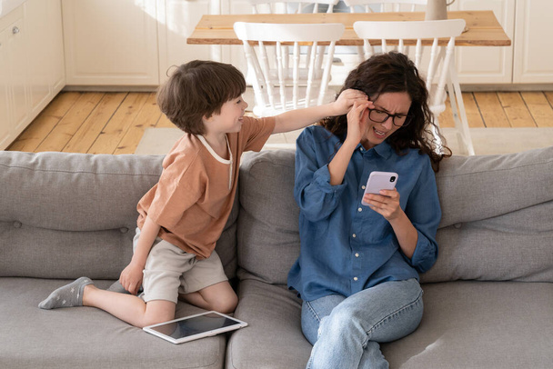 Jeune mère impuissante essayer de lire le message dans l'application smartphone assis sur le canapé avec un petit enfant désobéissant - Photo, image