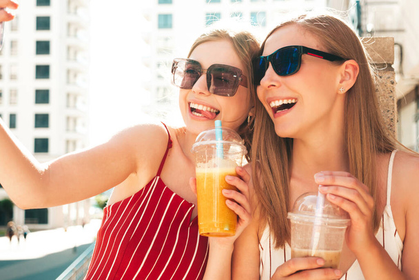 Two young beautiful smiling hipster female in trendy summer clothes. Carefree women posing outdoors.Positive models drinking fresh cocktail smoothie drink in plastic cup with straw.Taking selfie - Φωτογραφία, εικόνα