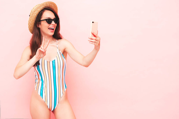 Young beautiful sexy smiling  hipster woman in posing near light blue wall in studio. Trendy model in colorful summer swimwear bathing suit. Positive model taking selfie.Happy and cheerful in hat - Foto, Imagem