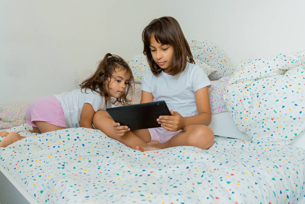 Two Caucasian sisters with their tablet in the bedroom sitting on their beds. - 写真・画像