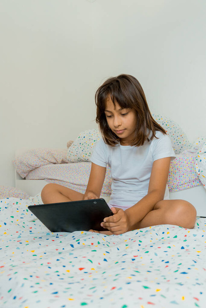 Běloch 9 letá dívka sedí na posteli ve svém pokoji a sleduje nebo hraje něco na svém tabletu. - Fotografie, Obrázek