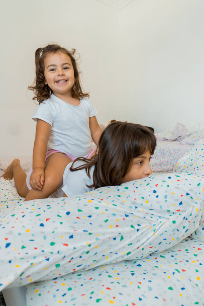 Zwei kaukasische Mädchen im Alter von zwei und neun Jahren amüsieren sich gemeinsam auf ihren Schlafzimmerbetten, spielen und reden. - Foto, Bild