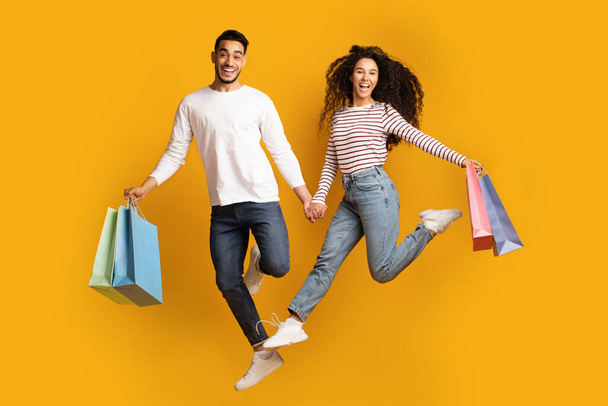 Cheerful Arabic Couple Holding Hands And Jumping With Shopping Bags - Photo, Image