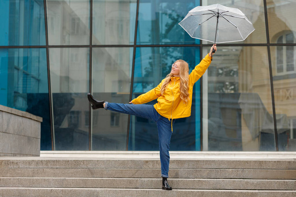 Szczęśliwa kobieta w żółtym płaszczu przeciwdeszczowym z parasolem na spacerze po mieście w deszczu - Zdjęcie, obraz