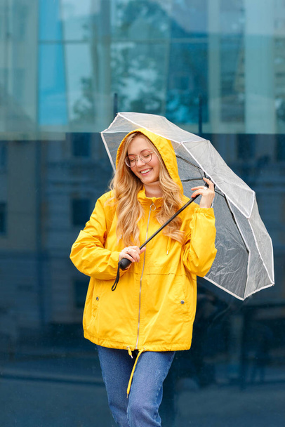 Frau lächelt in gelbem Regenmantel mit Regenschirm auf blauem Hintergrund in der Stadt - Foto, Bild