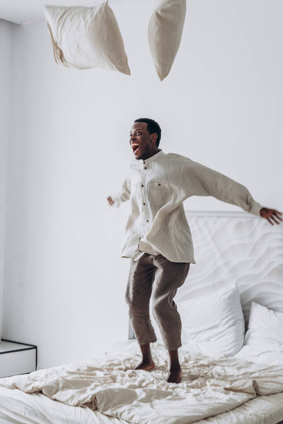 Joyful African-American man jumps throwing pillows on bed - Photo, Image