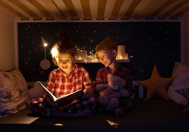 Cute girl smiling and reading fairytale to boy while sitting on bed near lamp in dark bedroom at night at home - Φωτογραφία, εικόνα