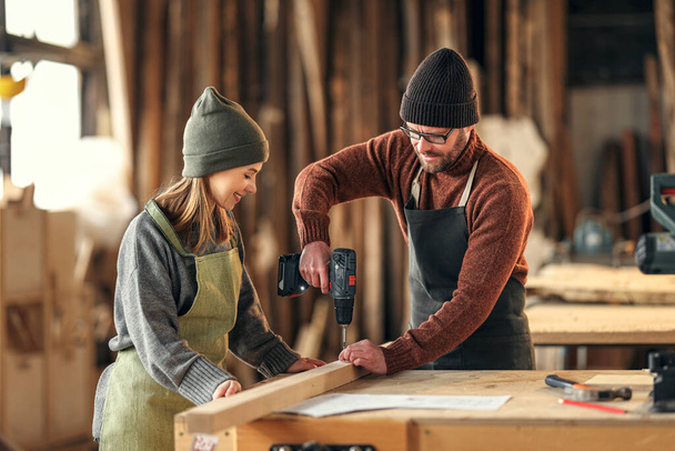 Positive young craftswoman assisting male partner drilling wooden plank with screwdriver while working together in carpentry workshop - Foto, Imagen