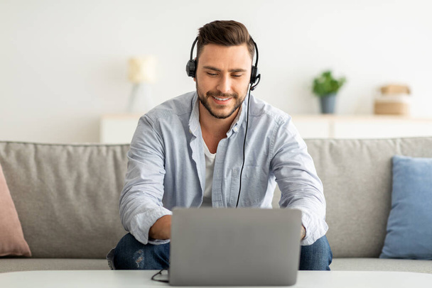 Concepto de aprendizaje a distancia. Hombre positivo en auriculares estudiando en línea a través de la computadora portátil, sentado en el sofá en casa, espacio de copia - Foto, imagen