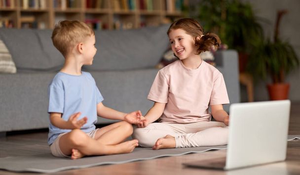 Yoga for kids. Two happy children, brother and sister in casual wear sitting in lotus pose in living room and meditating together with laptop looking at each other and smiling while watching tutorial online - Photo, Image