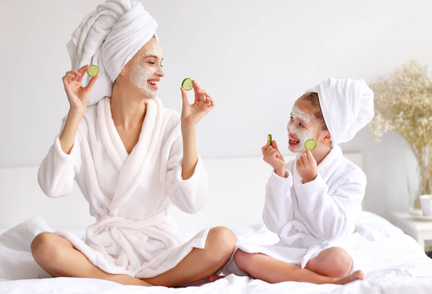 Jonge vrouw en meisje met komkommers glimlachen en kijken naar elkaar terwijl ontspannen op bed tijdens spa-procedure thuis - Foto, afbeelding
