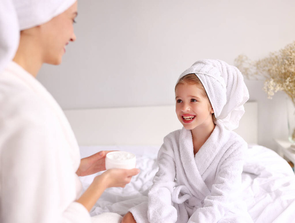 Crop woman applying cream on forehead of girl in bathrobe and towel sitting cross legged on bed during skin care routine at home - Photo, Image