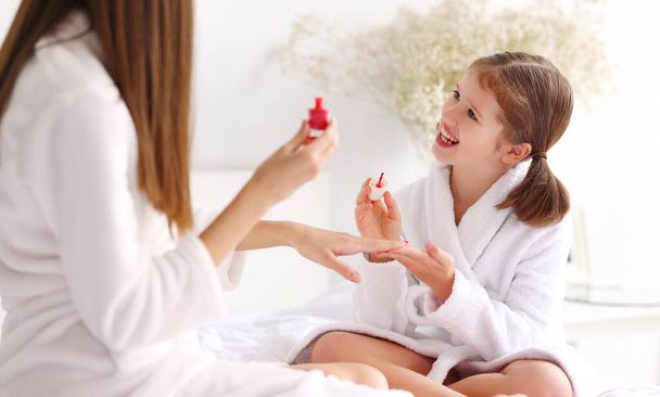 Cheerful girl in bathrobe smiling and doing manicure to woman while resting on bed during spa session at home - Photo, Image