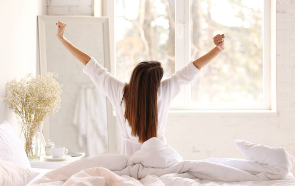 Πίσω όψη της ανώνυμης γυναίκας σε sleepwear σηκώνοντας τα χέρια και τεντώνοντας το σώμα, ενώ κάθεται στο κρεβάτι το πρωί στο υπνοδωμάτιο φως - Φωτογραφία, εικόνα