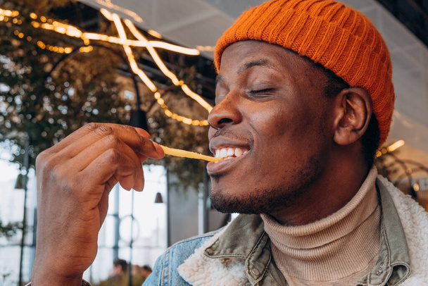 Black man in knitted hat enjoys french fries in cafe - Фото, зображення