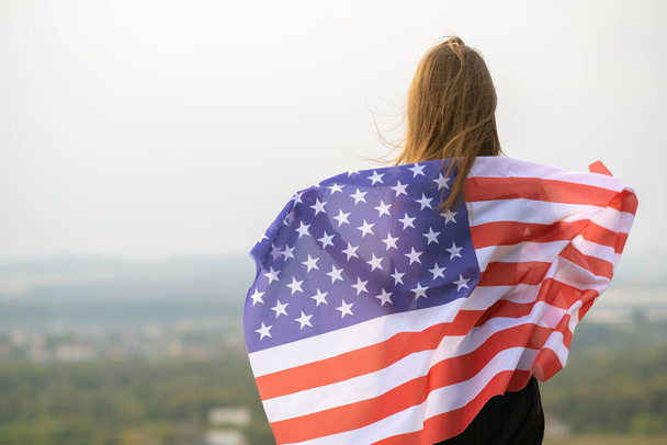 Young happy woman with long hair holding waving on wind american national flag on her sholders resting outdoors enjoying warm summer day. - Photo, Image