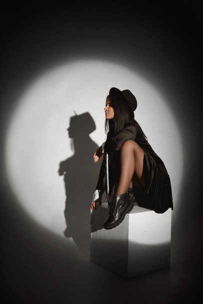 Fiatal modell lány fekete kalap és fekete ruha pózol egy fehér kocka helyszínen fény fehér háttér - Fotó, kép