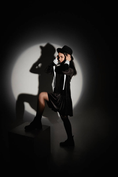 Молодая модель женщина в черной модной шляпе и короткое черное платье позируя в месте света в черной комнате - Фото, изображение