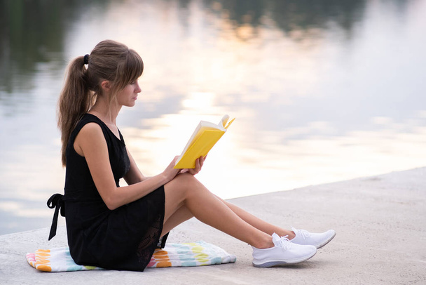 Female student sitting in summer park reading textbook outdoors. Education and sudy concept. - Фото, изображение