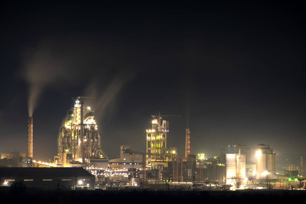 夜にタワークレーンや喫煙煙突とセメント工場の高いコンクリート構造物。工業生産と大気汚染の概念. - 写真・画像
