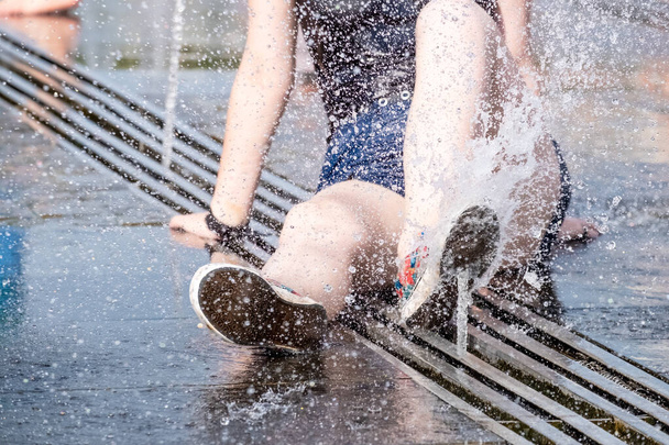 A young girl sits on the floor of a street fountain in splashes of water. - Photo, image