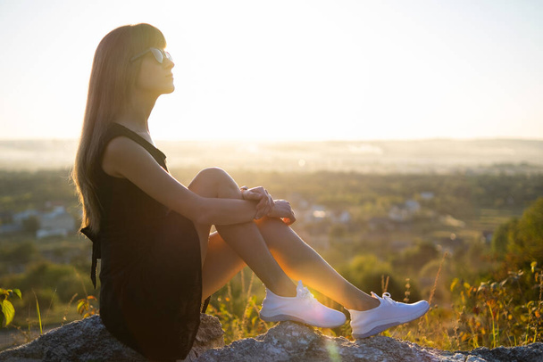 Joven mujer elegante en vestido corto negro sentado en una roca relajante al aire libre en la noche de verano. Mujer de moda disfrutando de la cálida puesta de sol en la naturaleza. - Foto, Imagen