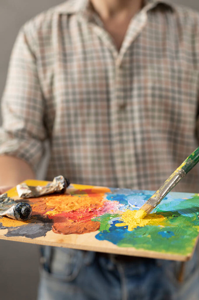 Man painter holding paint palette with paint brush. Young artist and paintbrush painting in creative studio - Fotó, kép