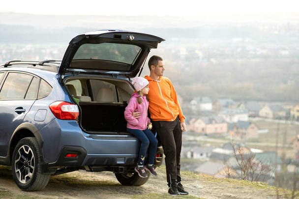 Side view of father with little daughter standing near car with open trunk and enjoying nature. Concept of weekend with family.  - Photo, Image