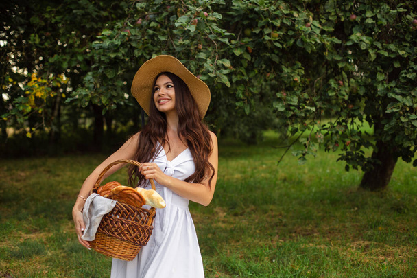白い歯を持つ若い美しい少女の肖像画、わらの帽子と長い白いドレスの美しい笑顔は庭にピクニックを持っています - 写真・画像