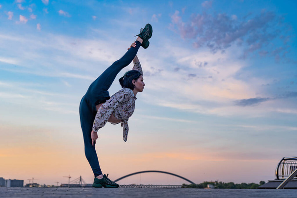 Silhouette of flexible yoga woman keeping balance in split on dramatic sunset and cityscape. Concept of healthy lifestyle and healthcare  - Photo, Image