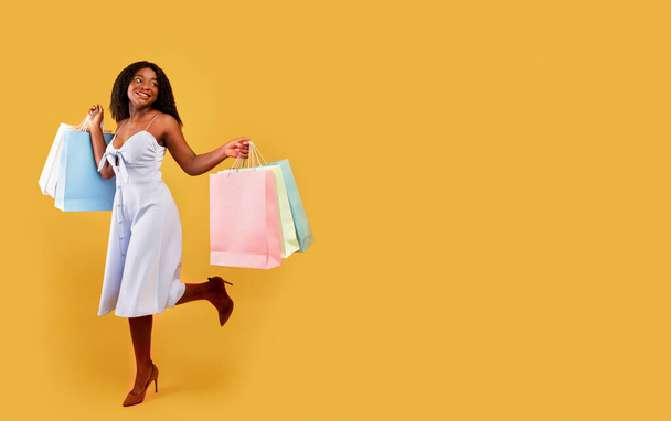 Longitud completa de la mujer negra despreocupada en vestido corriendo con bolsas de compras brillantes sobre fondo naranja, espacio libre - Foto, Imagen
