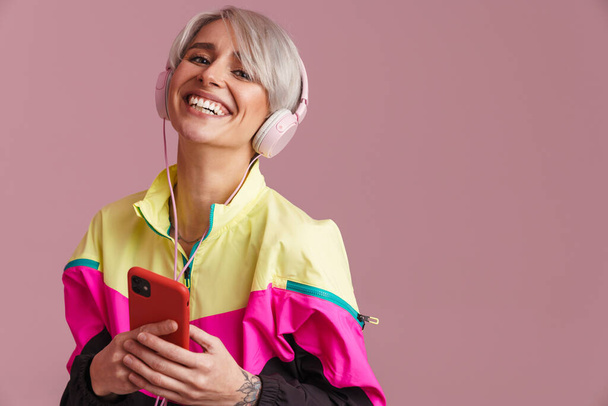 Mladá usměvavá žena poslech hudby se sluchátky a mobilní telefon izolované přes fialové pozadí - Fotografie, Obrázek