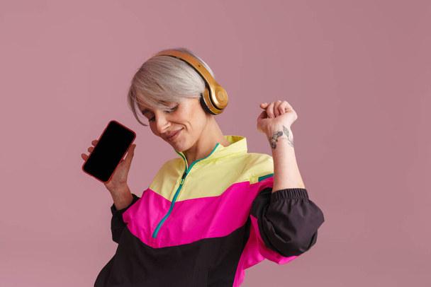 Usmívající se žena tanec při poslechu hudby se sluchátky a mobilní telefon izolované přes fialové pozadí - Fotografie, Obrázek