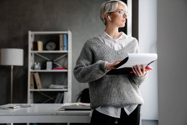 White-haired focused woman using earphone while examining documents in office - Foto, immagini