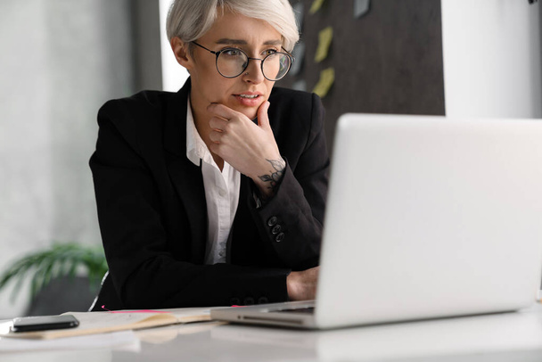 White-haired puzzled woman working with laptop while sitting at table in office - Foto, imagen