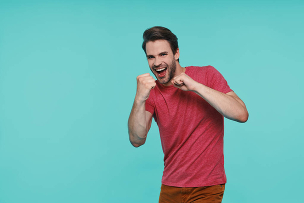 Playful young smiling man in casual clothing gesturing and smiling while standing against blue background - Foto, Bild