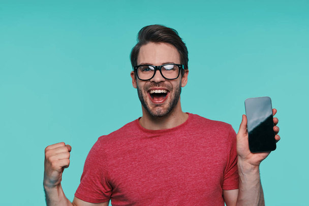 Portrait of handsome young smiling man in eyeglasses showing his smart phone and gesturing - Fotoğraf, Görsel