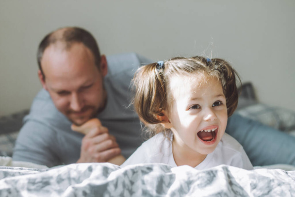 Apa játszik a kislányával 2-4-re az ágyban. Apa csiklandozza a gyerekek lábát a szakállával. A család jól érzi magát - Fotó, kép