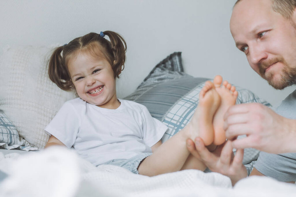 Ο πατέρας παίζει με την κορούλα 2-4 στο κρεβάτι. Ο μπαμπάς γαργαλάει τα πόδια των παιδιών. Η οικογένεια διασκεδάζει - Φωτογραφία, εικόνα