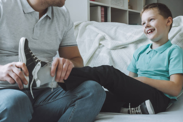 Dad and son sitting on couch. Father helps child to take off or put on shoes, kid laughs - Foto, immagini