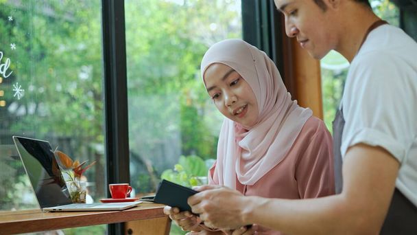 Selective focus, Young beautiful muslim woman wearing hijab typing on laptop, Waiter come in and She use credit card to payment in coffee shop - Φωτογραφία, εικόνα