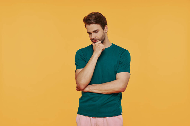Thoughtful young man in casual clothing holding hand on chin while standing against yellow background - Photo, image