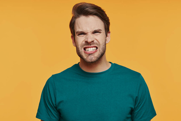 Portrait of angry young man looking at camera and making a face while standing against yellow background - Foto, afbeelding