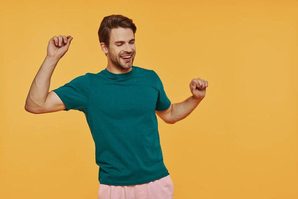 Handsome young man in casual wear dancing and smiling while standing against yellow background - Foto, Imagen