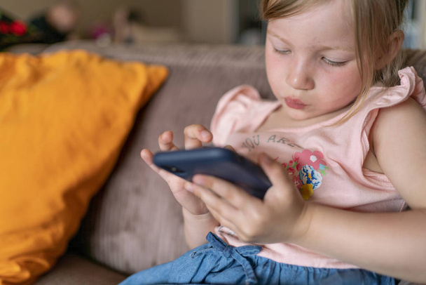 girl with blond hair plays with a smartphone at home. child and smartphone in hands. children with digital tablet - 写真・画像