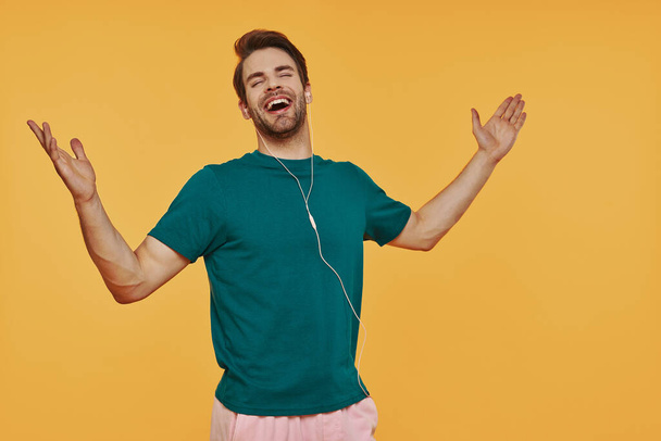 Красивий молодий чоловік в навушниках жестикулює і посміхається, стоячи на жовтому тлі
 - Фото, зображення